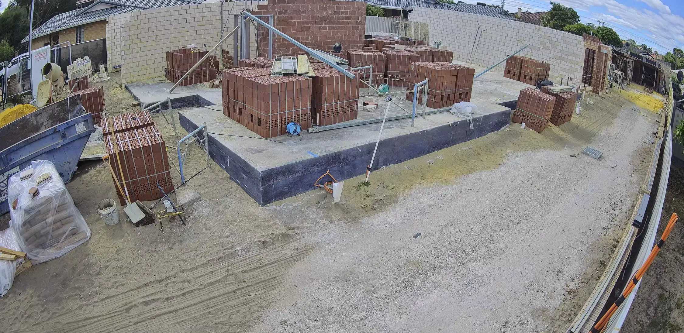Camera view across the concrete slab of a house under construction, with brick piers and stacked bricks laid out on it.